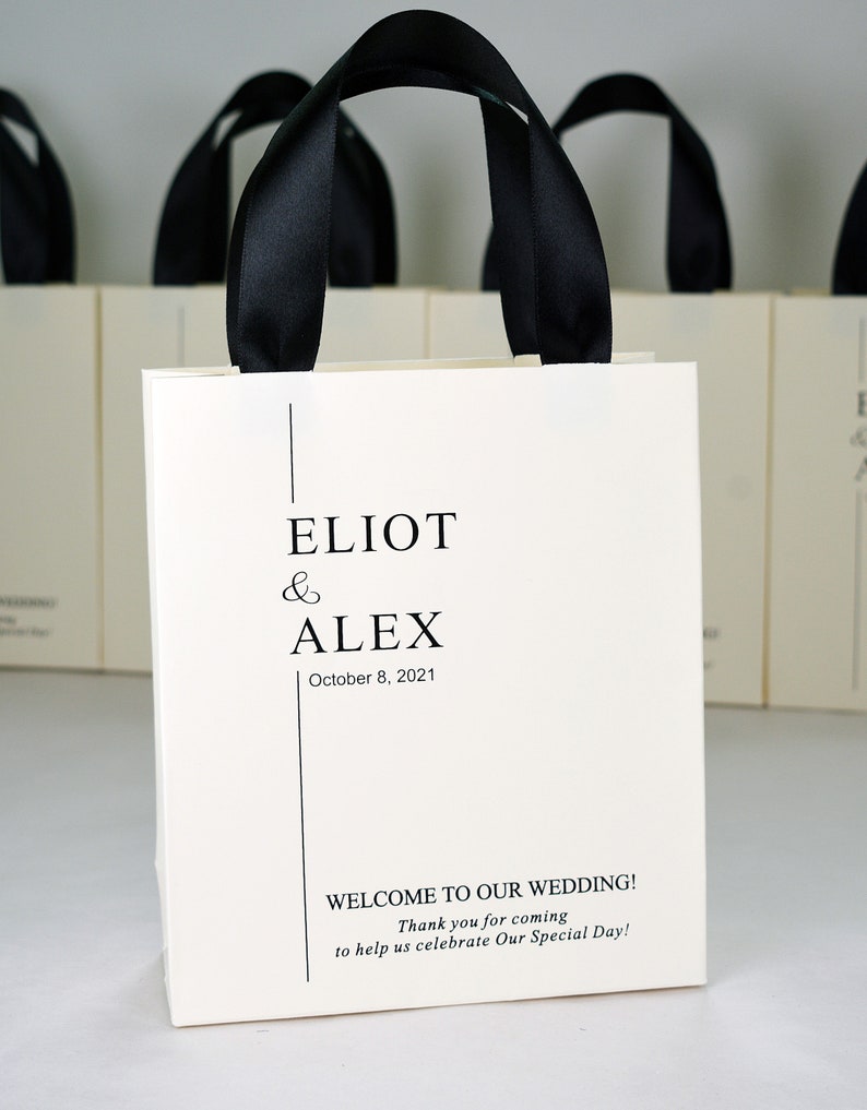 K&ouml;nnte beinhalten: Wei&szlig;e Papiert&uuml;te mit schwarzen Henkeln. Die Tasche tr&auml;gt den Text "ELIOT & ALEX" mit dem Datum "8. Oktober 2021" und die Nachricht "WILLKOMMEN ZU UNSERER HOCHZEIT! Vielen Dank, dass Sie gekommen sind, um mit uns unseren besonderen Tag zu feiern!"