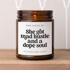 May include: A brown glass candle with a white label. The label has black text that reads 'WINK CANDLE CO.', 'She got mad hustle and a dope soul', '9OZ | SWEET BERRY VANILLA'.