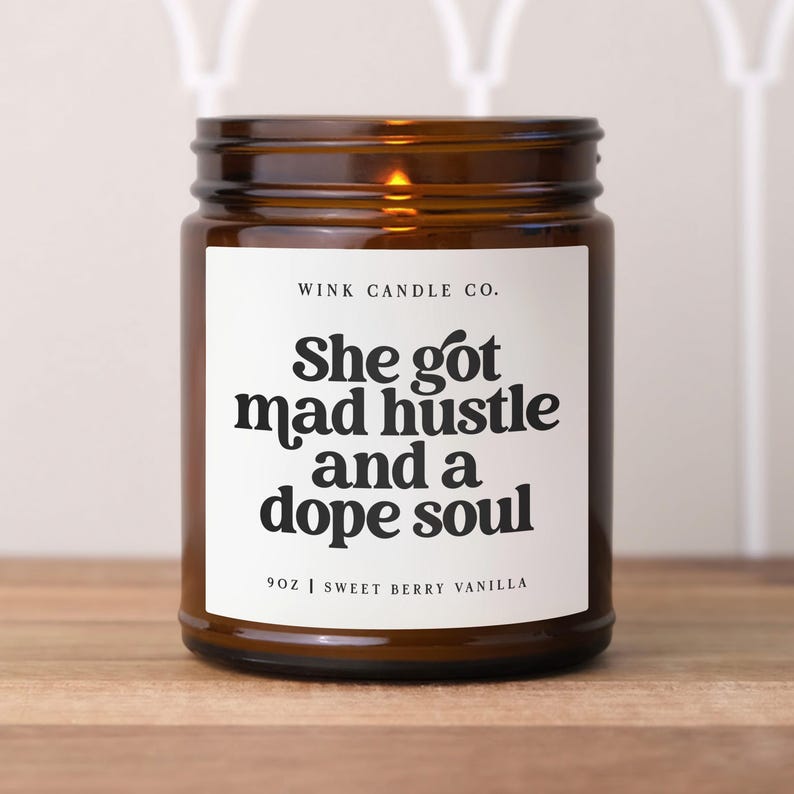 May include: A brown glass candle with a white label. The label has black text that reads 'WINK CANDLE CO.', 'She got mad hustle and a dope soul', '9OZ | SWEET BERRY VANILLA'.