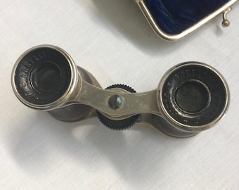 May include: Silver opera glasses with black leather accents and a focus wheel. The glasses have the words "GILBERT" and "CHICAGO" printed on the lenses.