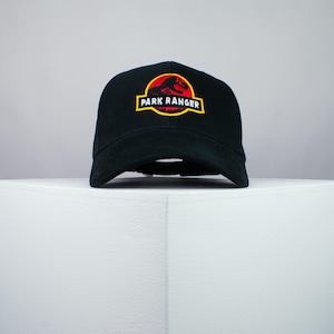 May include: Black baseball cap with a curved brim. The cap features a circular patch with a red and yellow design and the words "PARK RANGER" in a yellow rectangle.