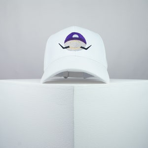 Puede incluir: Gorra de béisbol blanca con un diseño bordado en morado y dorado de un personaje con bigote. El personaje lleva un sombrero morado.