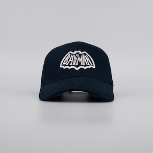 Può includere: Un cappellino da baseball blu navy con il logo di Batman ricamato in bianco. Il logo è una silhouette stilizzata di pipistrello con la scritta "Batman" sopra. Il cappellino ha una visiera curva e un classico design a sei pannelli.