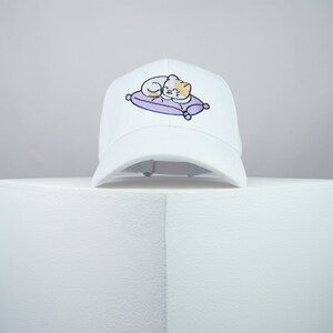 May include: White baseball cap with an embroidered design of a cat sleeping on a purple cushion.
