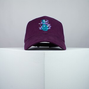 May include: A purple baseball cap with an embroidered design of a blue bottle with purple smoke and white stars.