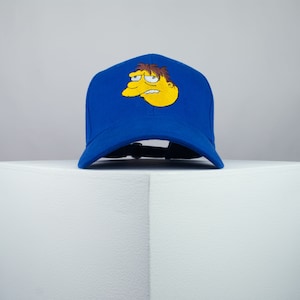 May include: A blue baseball cap with a yellow cartoon character embroidered on the front. The character has brown hair and a yellow face with a frown.