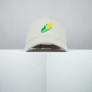 Può includere: Un berretto da baseball bianco con una pannocchia di mais gialla ricamata con foglie verdi.