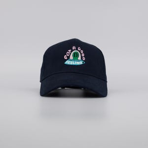 Può includere: Cappellino da baseball blu navy con visiera curva. Il cappellino presenta un design ricamato con le parole "GOT A GOOD FEELING" in rosa e azzurro, che circondano un cactus verde.