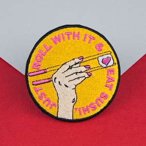 May include: Embroidered patch with a yellow circular background and black border. The patch features a hand holding sushi with pink chopsticks. The text "Just roll with it & eat sushi" is in pink.