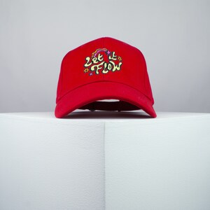 May include: Red baseball cap with an embroidered design that says "Let It Flow" in yellow and pink lettering with stars.