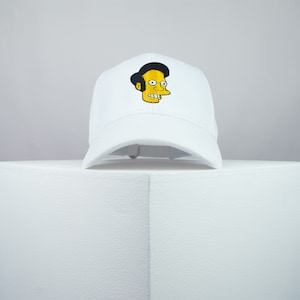 May include: White baseball cap with an embroidered cartoon character with yellow skin, black hair, and a wide smile.