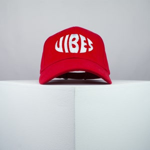 Puede incluir: Gorra de béisbol roja con un logotipo blanco "VIBES" en una fuente redondeada.