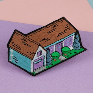 Puede incluir: Parche bordado para planchar de una casa azul y blanca con techo marrón, puerta y ventanas moradas y arbustos verdes.