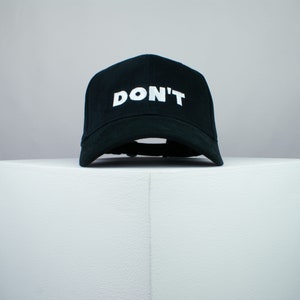 Könnte beinhalten: Eine schwarze Baseballkappe mit einem weißen, gestickten "DON'T" auf der Vorderseite.