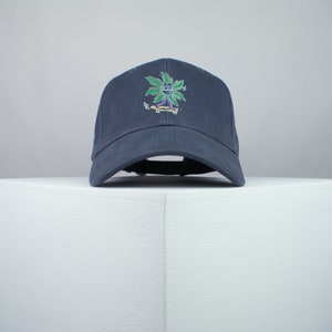 May include: A dark blue baseball cap with an embroidered green marijuana leaf character skateboarding. The character has a purple face and yellow skateboard.