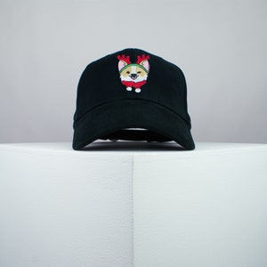 May include: Black baseball cap featuring an embroidered corgi wearing a reindeer antler headband and a red and white outfit. The cap is displayed on a white surface.
