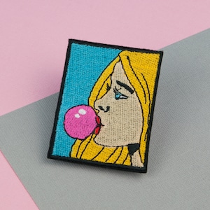 Könnte beinhalten: Gestickter Bügel-Patch mit einer Frau im Cartoon-Stil, die eine rosa Blase bläst. Die Frau hat blonde Haare und trägt eine schwarze Halskette.