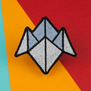 Op de afbeelding: Een witte en grijze origami-stijl papieren vliegtuig patch met zwarte stiksels. De patch is op een gele en rode achtergrond.