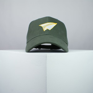 May include: A dark green baseball cap with a white paper airplane embroidered on the front. The airplane has a yellow outline.