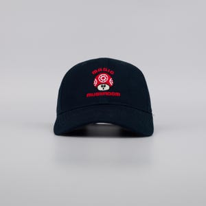 May include: A navy blue baseball cap with a red and white mushroom design. The word "MAGIC" is embroidered above the mushroom, and "MUSHROOM" is below. The cap has a curved brim and a classic six-panel design.