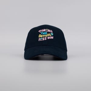 May include: A navy blue baseball cap with the text "TAKING THE ACTUAL PESS NOW" in colorful, playful lettering. The cap has a curved brim and a classic six-panel design, suitable for casual wear.