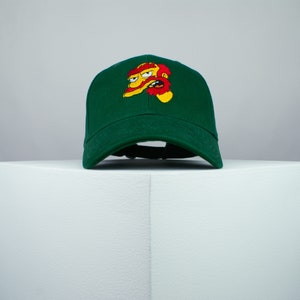 Op de afbeelding: Een groene baseballpet met een geborduurd cartoonfiguur op de voorkant. Het figuur heeft een geel gezicht, rood haar en een rode baard.