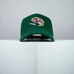 May include: A green baseball cap with an embroidered design of a yellow cloud with the words "OUR EARTH" in pink and black lettering.