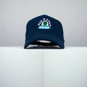 Può includere: Un cappello da baseball blu navy con un design ricamato di un UFO con il testo "Got a good feeling" in rosa e blu.