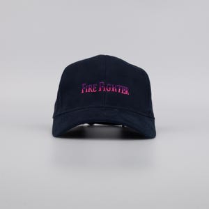 May include: A navy blue baseball cap with the embroidered text "FIRE FIGHTER" in a gradient pink color. The cap has a curved brim and a classic six-panel design.