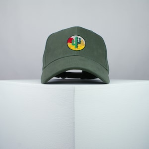 May include: A dark green baseball cap with a round embroidered patch featuring a cactus and a sunset. The patch is yellow, red, and green.