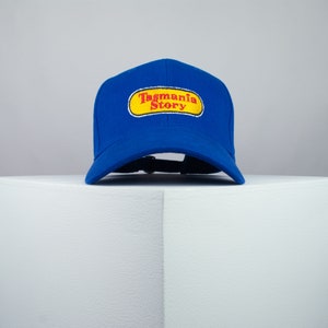 May include: A royal blue baseball cap with a yellow oval patch that says "Tagmania Story" in red lettering.
