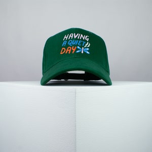 May include: A green baseball cap with the text "HAVING A QUIET DAY" embroidered in pink, orange, and blue. A blue and white dragonfly is embroidered next to the text.