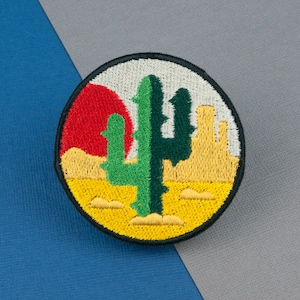 May include: A round embroidered patch featuring a green cactus in the centre, a red sun setting behind it, and yellow sand dunes in the background.