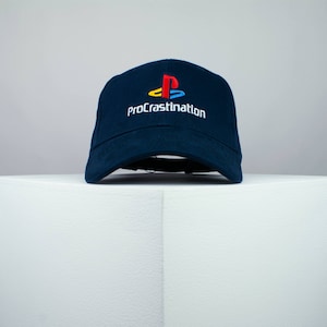 May include: A dark blue baseball cap with a white embroidered PlayStation logo and the text "proCrastination".