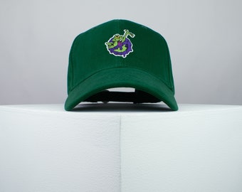 Op de afbeelding: Een donkergroene baseballpet met een geborduurd cartoonfiguur op de voorkant. Het figuur is groen en paars en lijkt een monster te zijn.