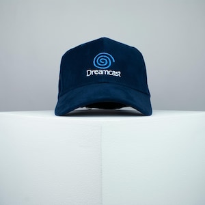 May include: A navy blue baseball cap with a white embroidered Dreamcast logo featuring a blue swirl.