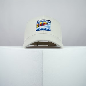 May include: A white baseball cap with an embroidered patch featuring a blue and white seascape with houses and the text "AMSTERDAM".