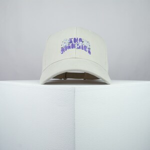 Puede incluir: Una gorra de béisbol beige con un diseño bordado en morado que dice "The Highlife" con flores blancas alrededor del texto.