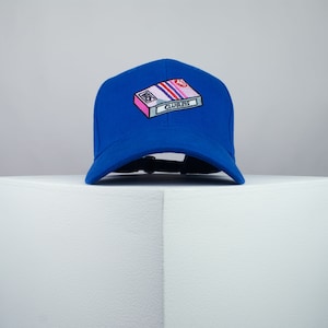 Puede incluir: Una gorra de béisbol azul con un diseño bordado de cinta VHS blanca y rosa que dice "Clueless" en ella.