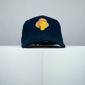 May include: A dark blue baseball cap with an embroidered image of a cartoon character with yellow skin, brown hair, and a blue earring. The character is wearing a pink dress and has a surprised expression.