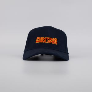 May include: A navy blue baseball cap with the words "FIGHTING SIMULATOR" embroidered in red and yellow. The cap has a curved brim and a structured crown. This hat is a great accessory for casual wear.
