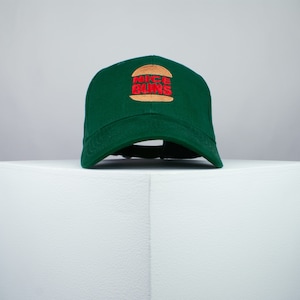 Pode incluir: Um boné de beisebol verde com um gráfico de hambúrguer marrom e amarelo e o texto "NICE BUNS" em vermelho.