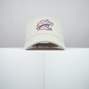 May include: A beige baseball cap with a pink and blue embroidered patch that says "Sydney" with palm trees.