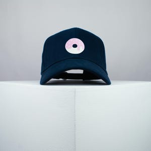 以下が含まれることがあります： ピンクのフロスティングをかけた白いドーナツのグラフィックが施されたネイビーブルーのベースボールキャップ