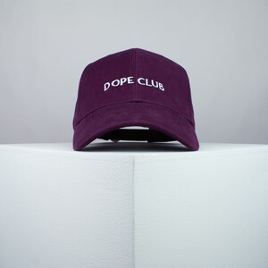 Może przedstawiać: Fioletowa czapka z daszkiem z haftowanym białym napisem "DOPE CLUB".
