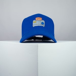 May include: A blue baseball cap with a white and grey embroidered image of a toaster with bread popping up.