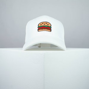 Op de afbeelding: Witte baseballpet met een geborduurd beeld van een hamburger op de voorkant.