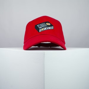 May include: Red baseball cap with a black and white embroidered image of a vintage VHS cassette tape with the text "E.T."