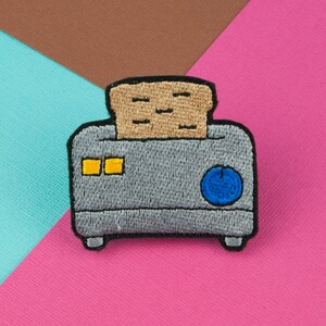 Könnte beinhalten: Ein grauer, gestickter Toaster mit einer braunen, gestickten Scheibe Brot darin. Der Toaster hat einen blauen Knopf und zwei gelbe Quadrate an der Seite.