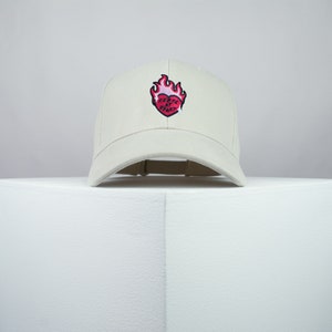 May include: A white baseball cap with a red heart with flames on it. The heart has the text "Keep it by heart" on it.